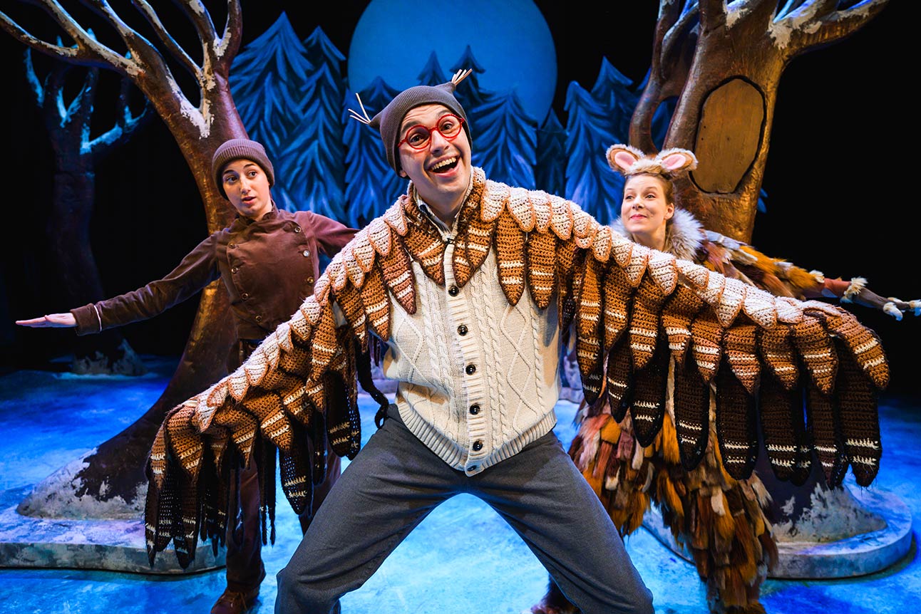 Three actors in whimsical costumes perform on stage, with a large blue moon and trees behind them.
