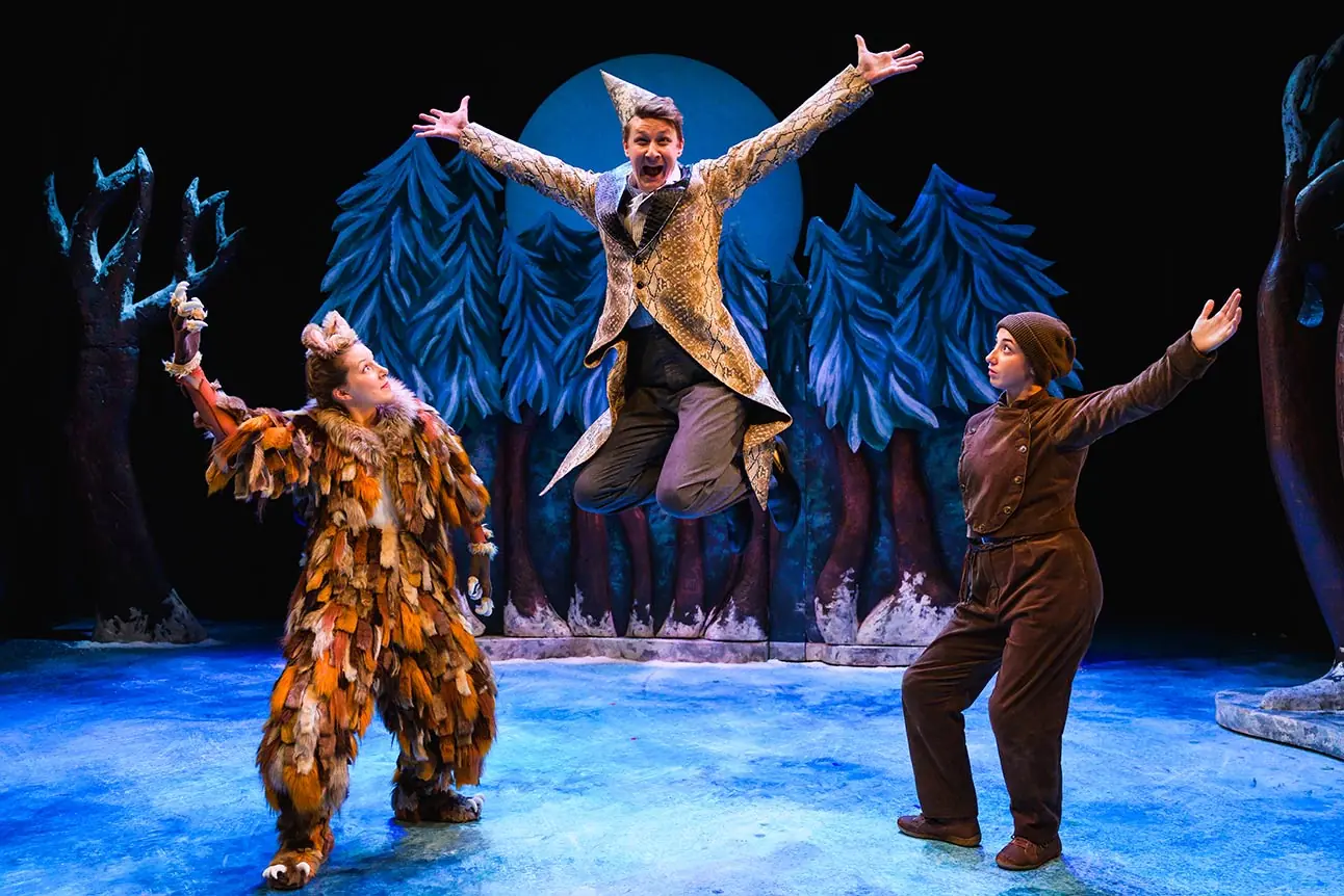 Three actors in vibrant costumes perform on stage, surrounded by blue trees and a dark backdrop.