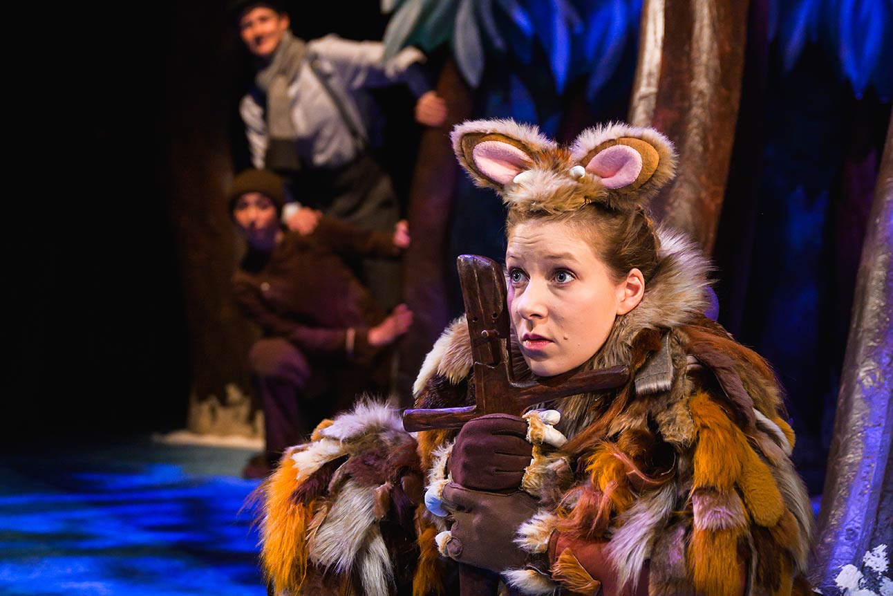 A young actress in a brown furry costume holds a wooden prop, with trees in the background.