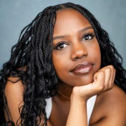 A woman with braided hair rests her chin on her hand, wearing a white top, grey background.
