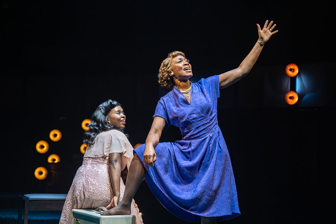 Two women perform on stage; one in a blue dress sings, the other watches in pink.