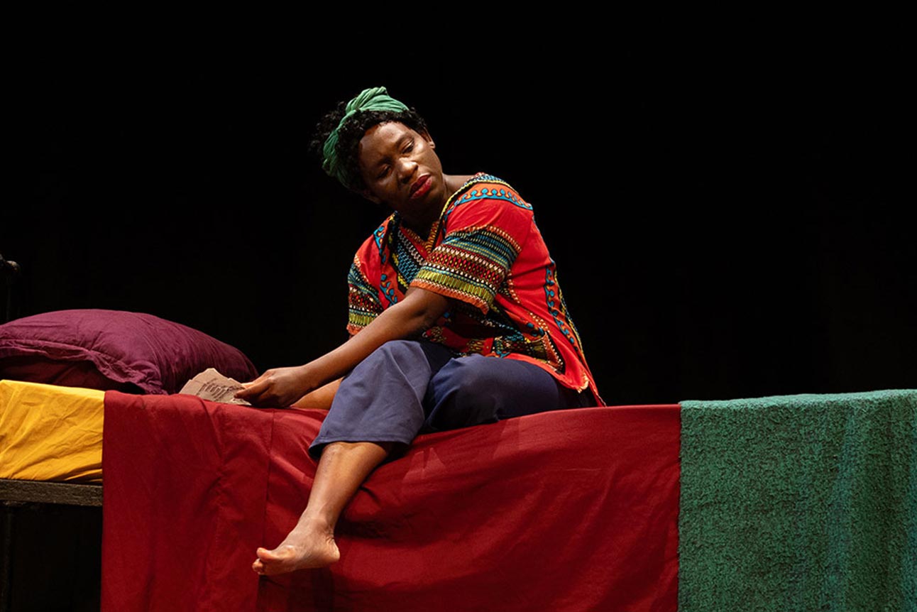 A person in a red patterned top and green headwrap sits barefoot on a bed with colourful linens.