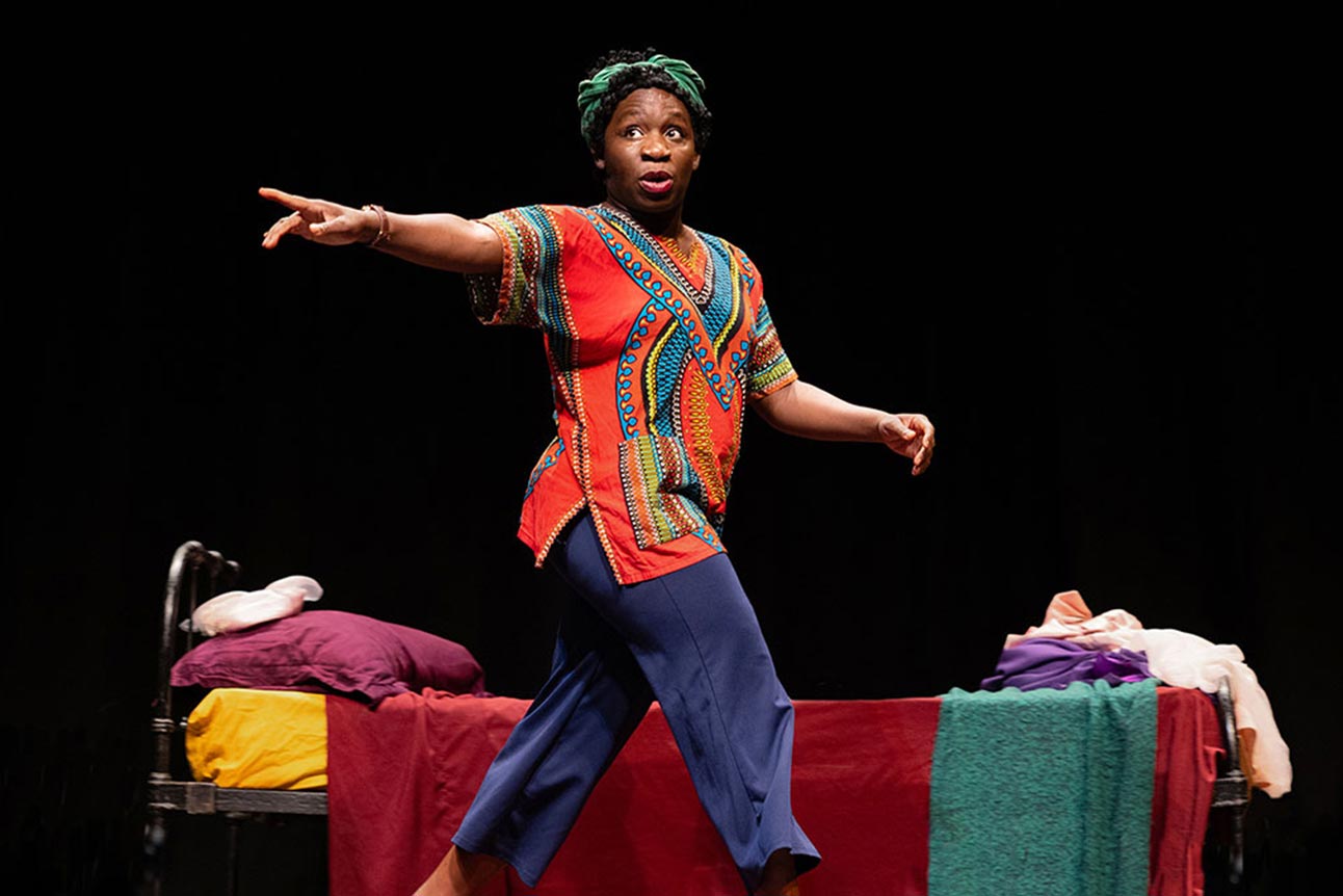 A person in a colourful shirt, performing on stage with a bed in the background.