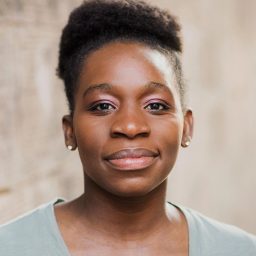 Confident Black woman with natural textured hair, wearing light green top and subtle pink eyeshadow, neutral background.