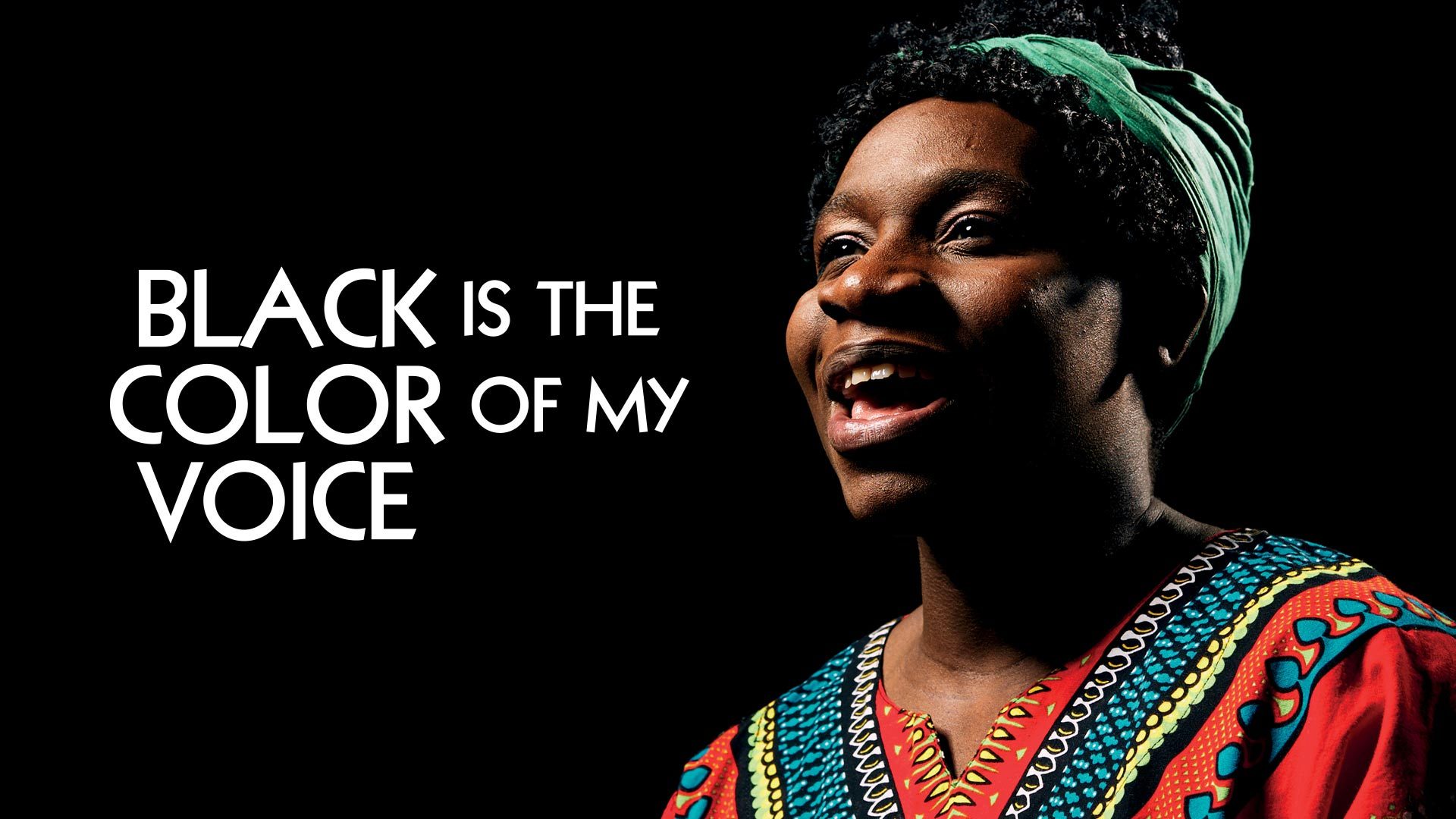 A person in a vibrant patterned shirt, smiling against a dark background.