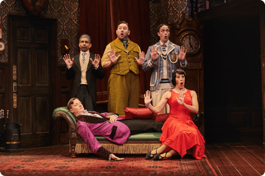 Five actors in vibrant costumes express shock; one reclines on a green sofa with ornate wallpaper.