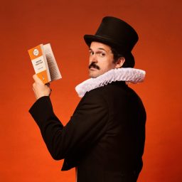 Garry Starr dressed in a black tuxedo with white ruff and top hat, holding a book against an orange background.