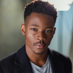 Alistair Nwachukwu wearing a dark suit and white shirt, smiling against a plain grey background.