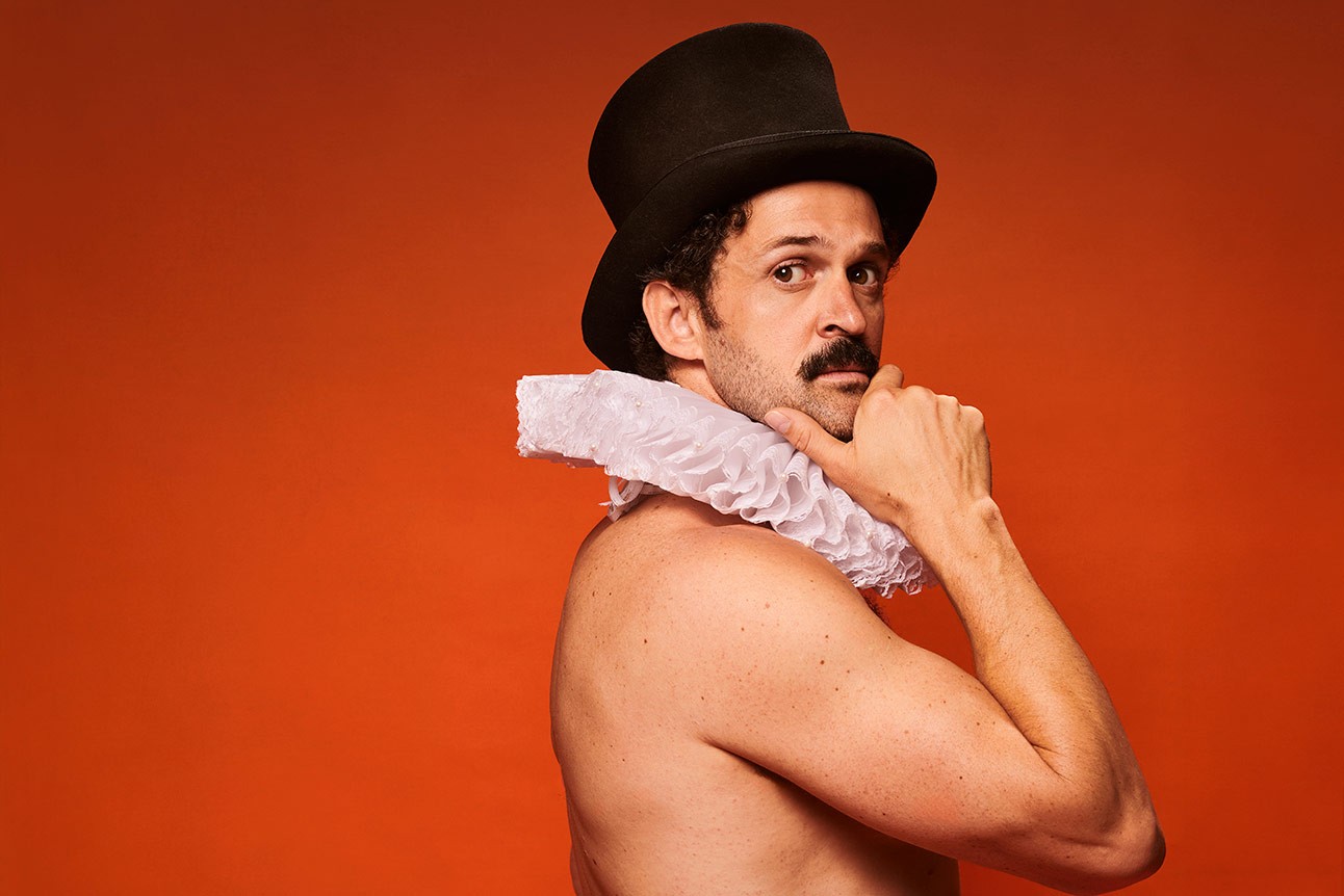 Garry Starr shirtless wearing a black top hat and white ruffled collar against an orange background.