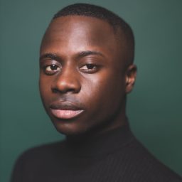 Idriss Kargbo wearing a black turtleneck, looking directly at the camera against a green background.