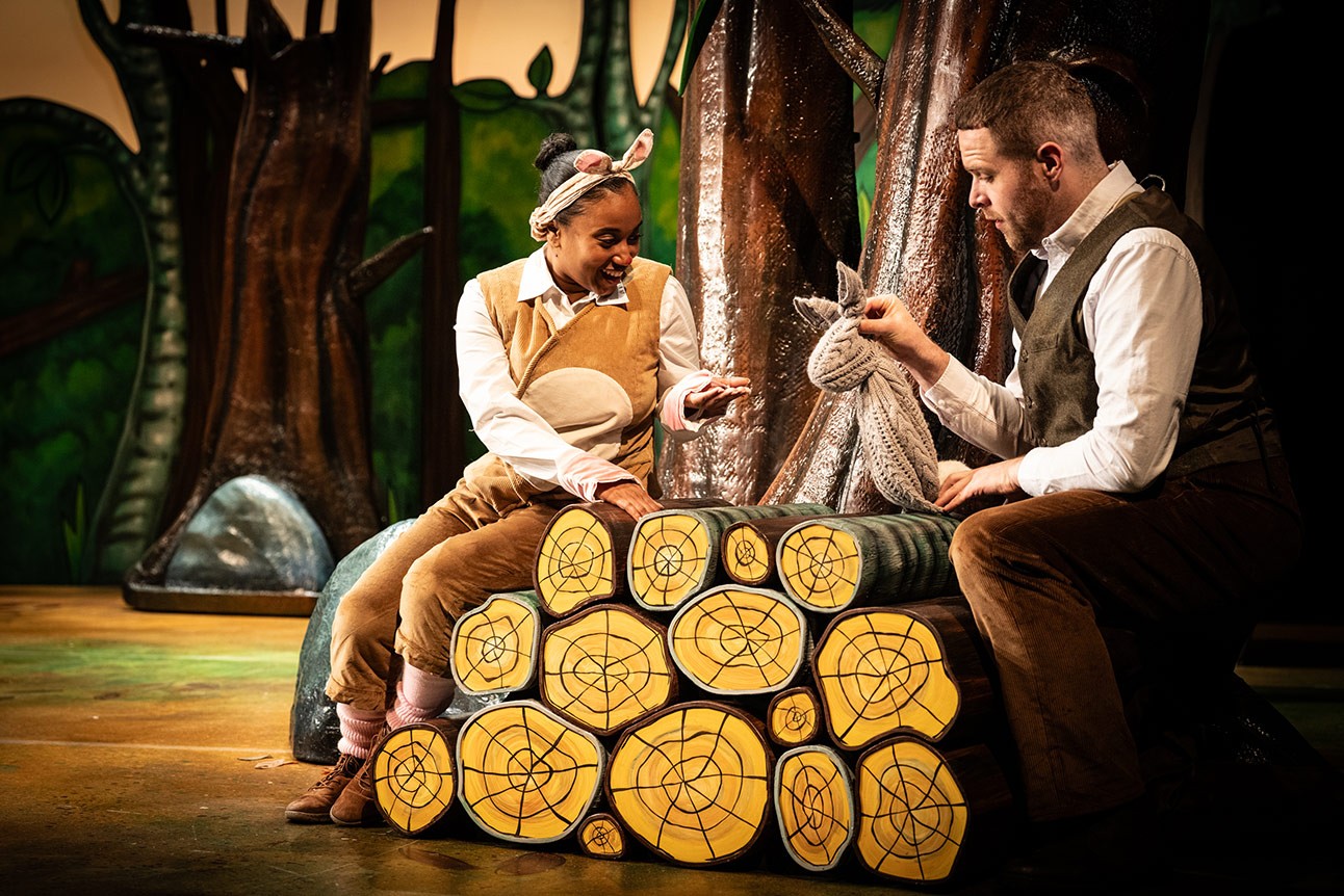 Rianna Kellman and Niall Hemingway perform on stage, sitting on log prop, vibrant forest backdrop.