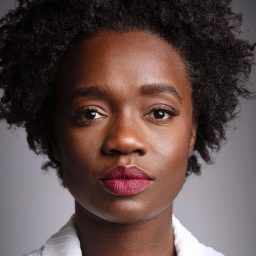 Madeline Appiah smiles against a plain grey background, wearing a blue top with natural curly hair.