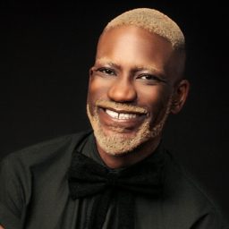 Newtion Matthews wearing a dark suit and white shirt, smiling against a plain grey background.