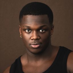 Owen Chaponda wearing a dark jacket and light shirt, smiling against a plain grey background.