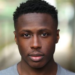 Shaka Kalokoh smiles in a close-up headshot with a neutral grey background, wearing a dark shirt.