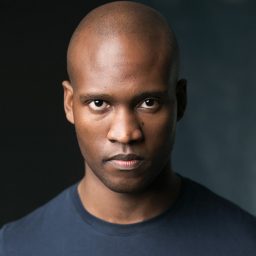 Sifiso Mazibuko wearing a dark suit and white shirt, smiling against a plain grey background.