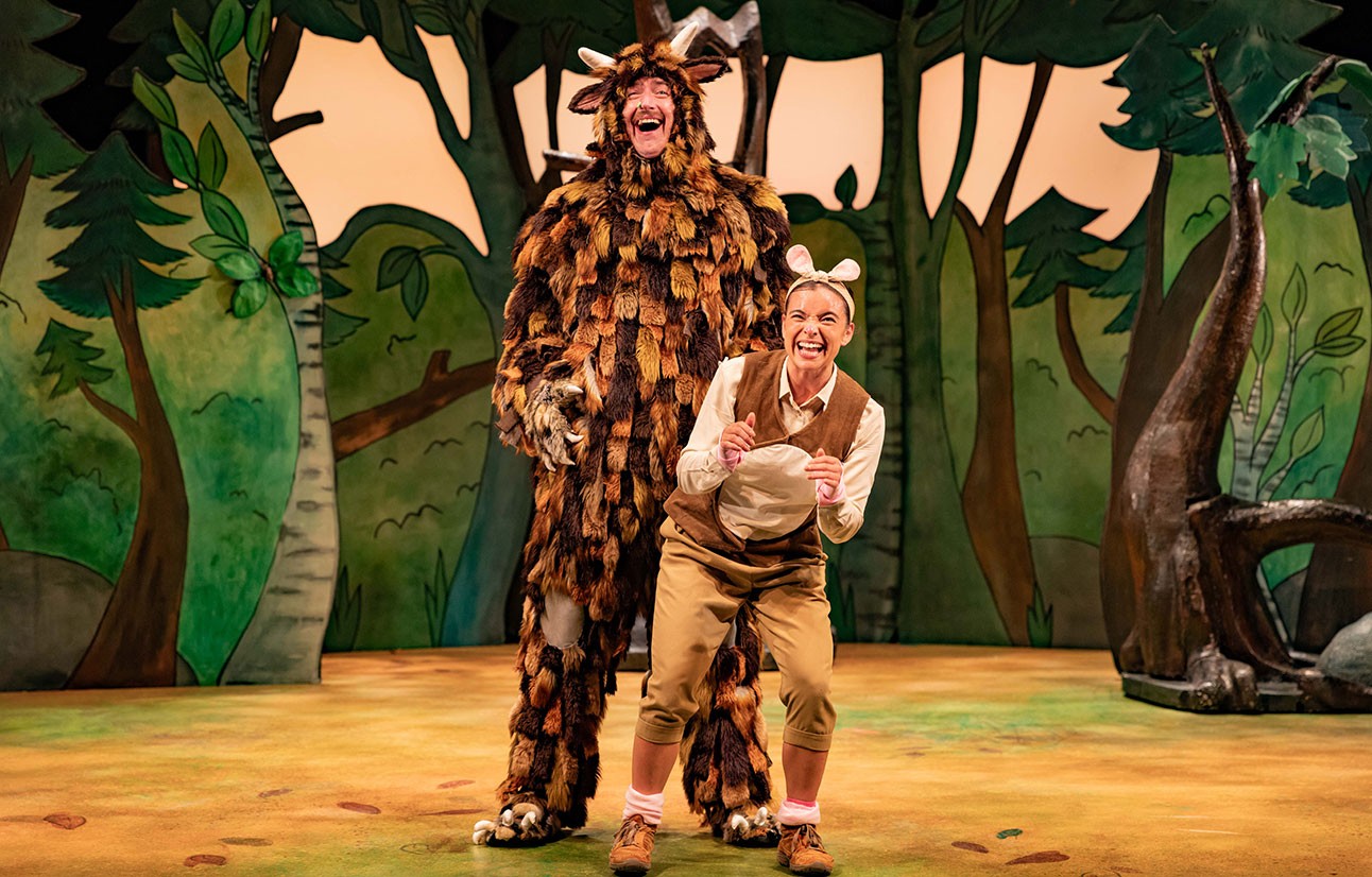 Actor in a Gruffalo costume with brown fur and orange eyes on a forest stage background.