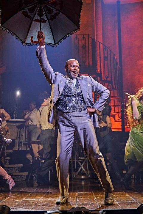 Cedric Neal in costume singing on stage with dark background and warm, dramatic lighting in Hadestown.