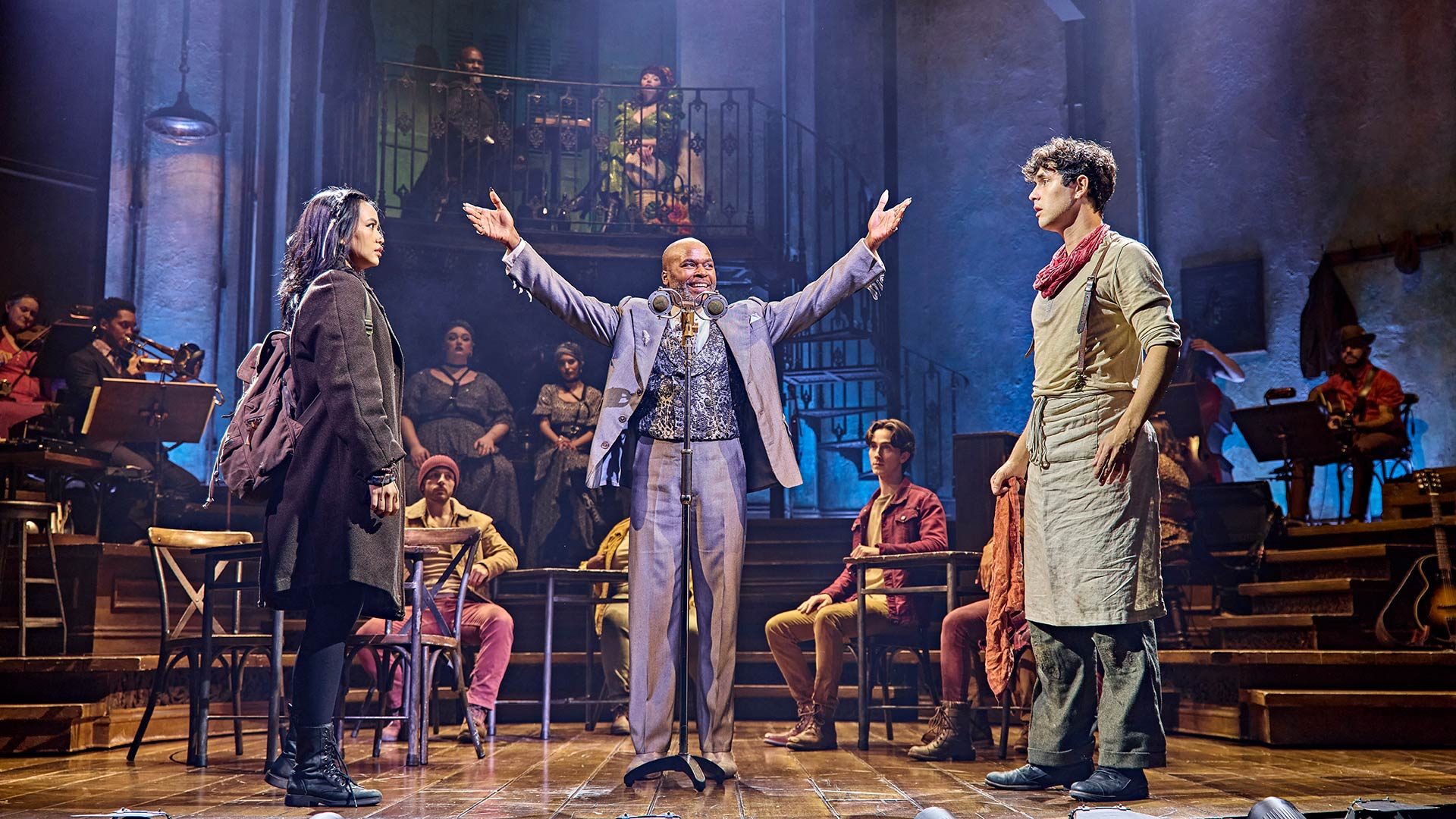 Hadestown Desmonda Cathabel, Cedric Neal, and Dylan Wood perform on stage in colourful costumes with a dark theatrical backdrop.