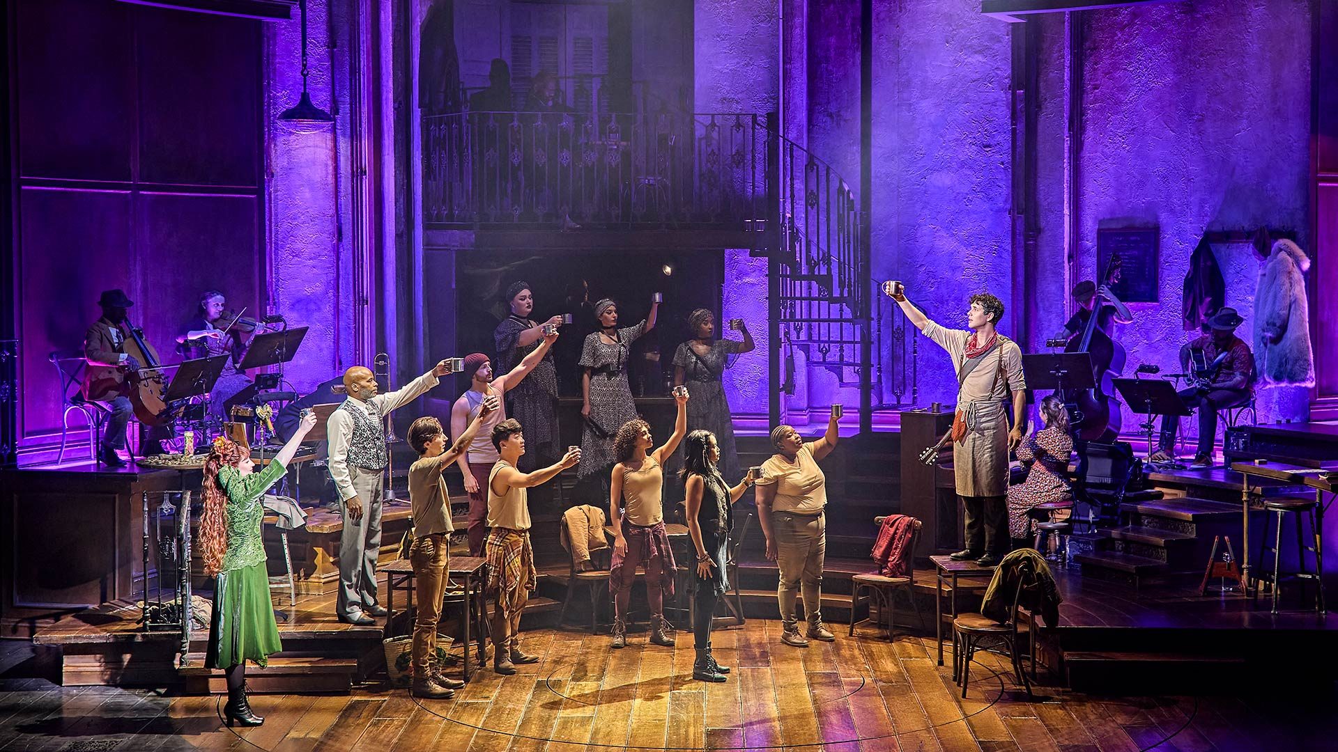 Hadestown The Hadestown cast stands in colourful costumes on stage, smiling against a dark theatre backdrop.