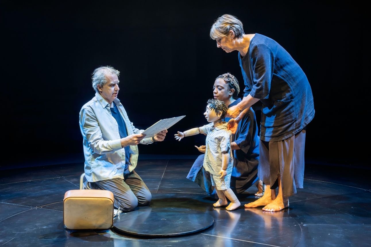 A man kneels holding a Philip Glass album, facing a woman and child puppet on a dimly lit stage.