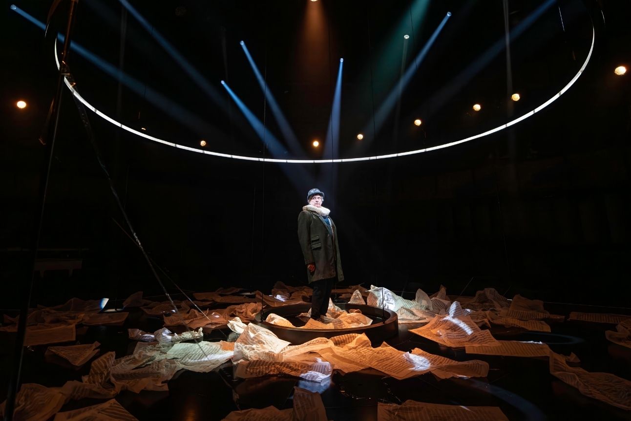 A person stands centre stage under dramatic lighting, surrounded by scattered sheet music paper.