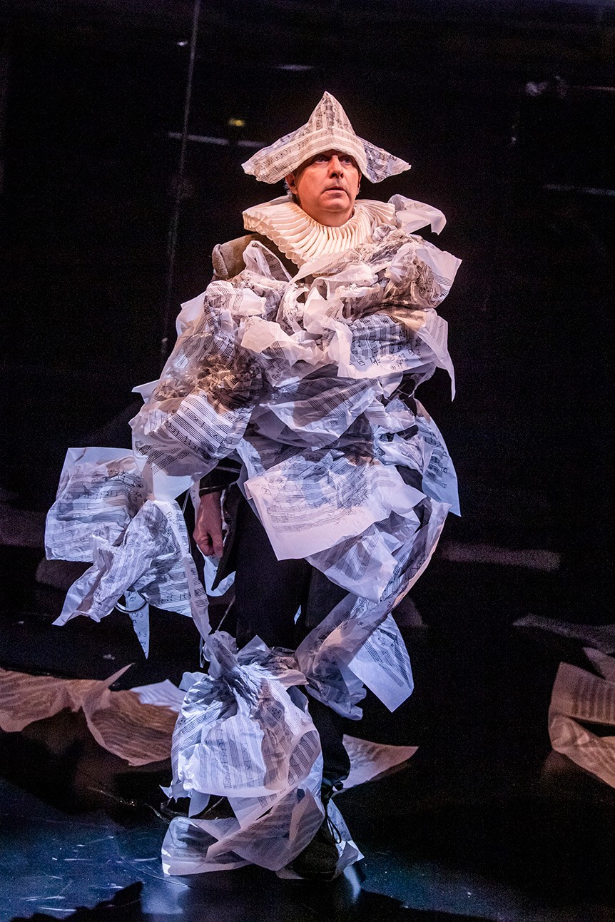Phelim McDermott in costume covered with sheet music, performing on stage, dark background.
