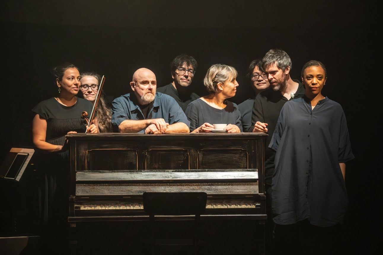 The musicians and puppeteers of Tao of Glass stand behind a wooden piano, dimly lit, wearing dark clothing.