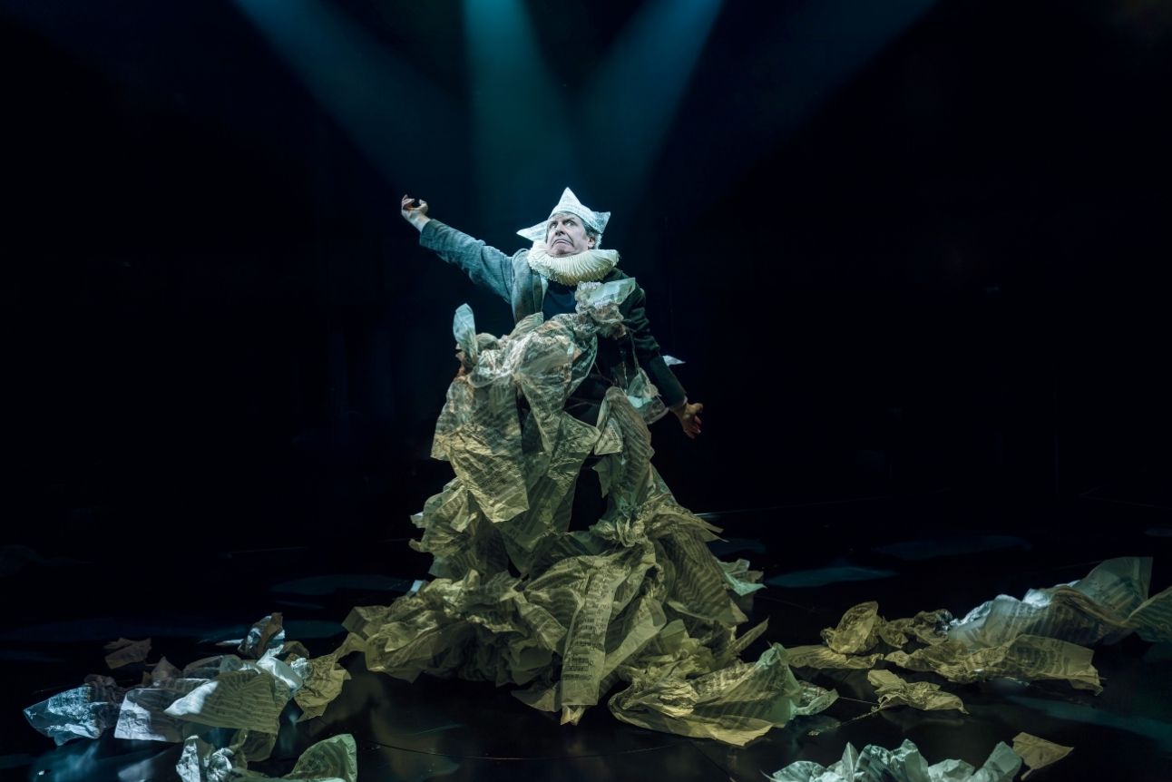 Phelim McDermott stands centre stage wearing a paper hat, reaching out to the side, with dramatic lighting and surrounded by crumpled sheet music paper.