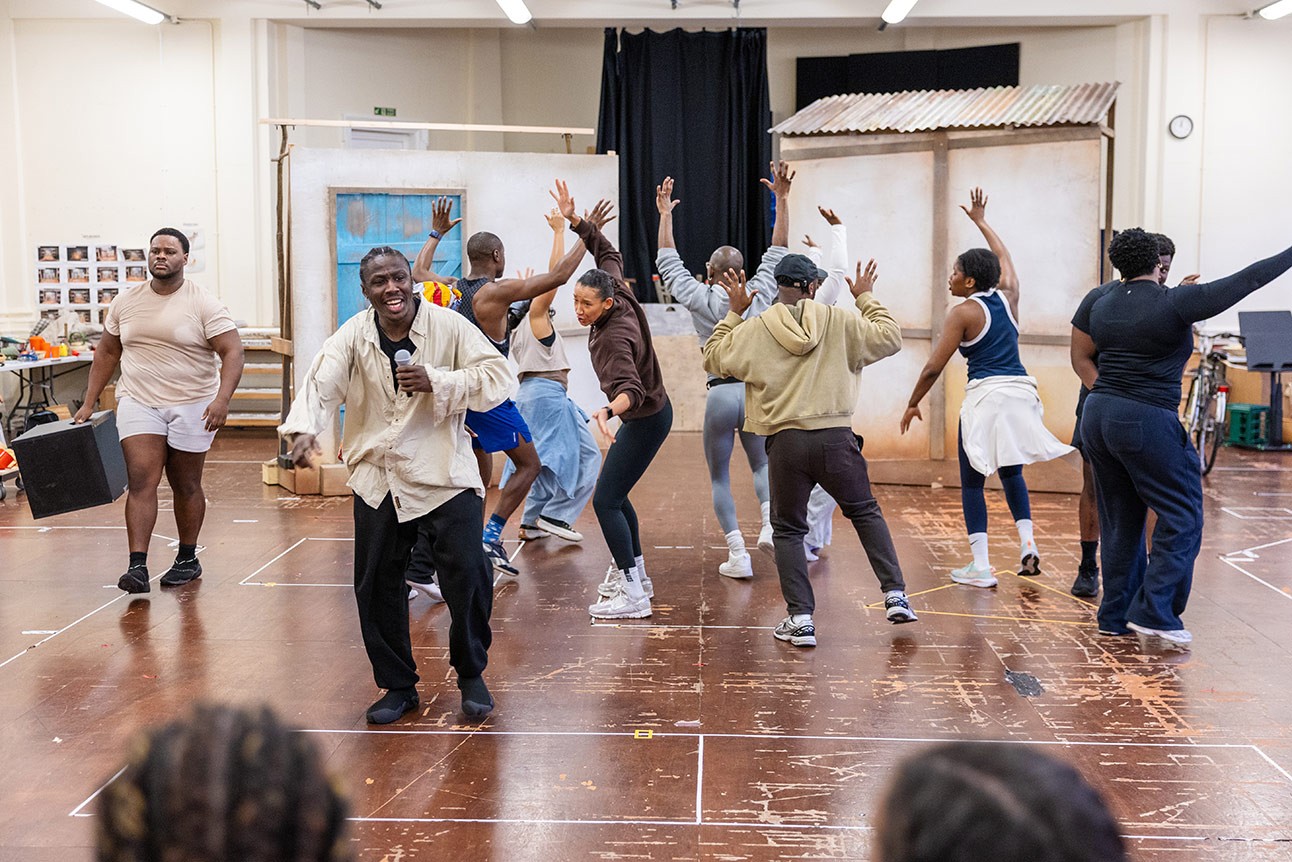 Actors rehearsing a lively dance scene on stage with casual clothing and a simple indoor background.