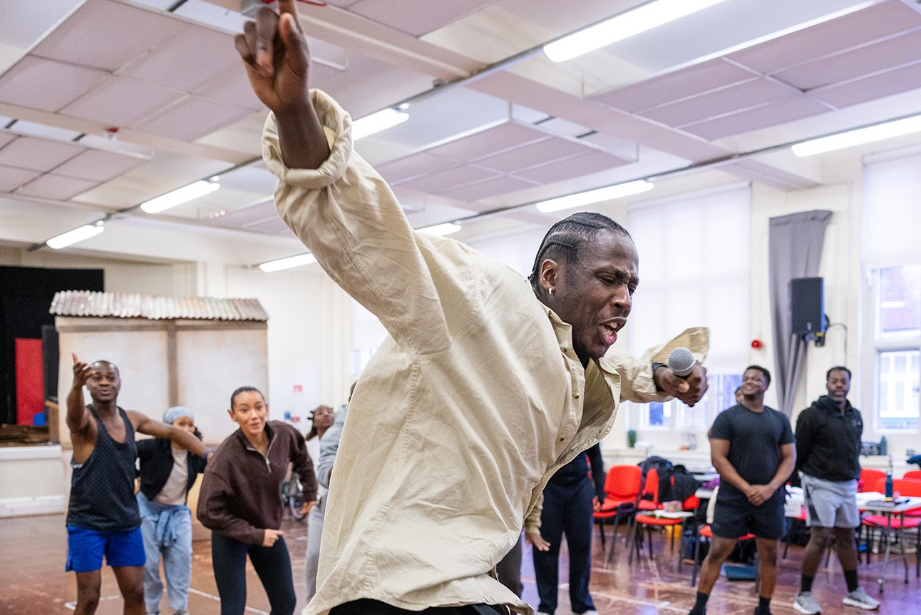 Young man in beige shirt gestures energetically during rehearsal, with standing people in background.
