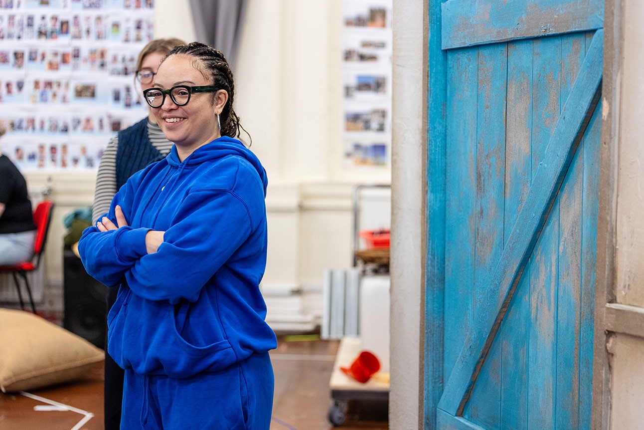 Woman in blue tracksuit and glasses stands with arms crossed, smiling, near a blue wooden door indoors.