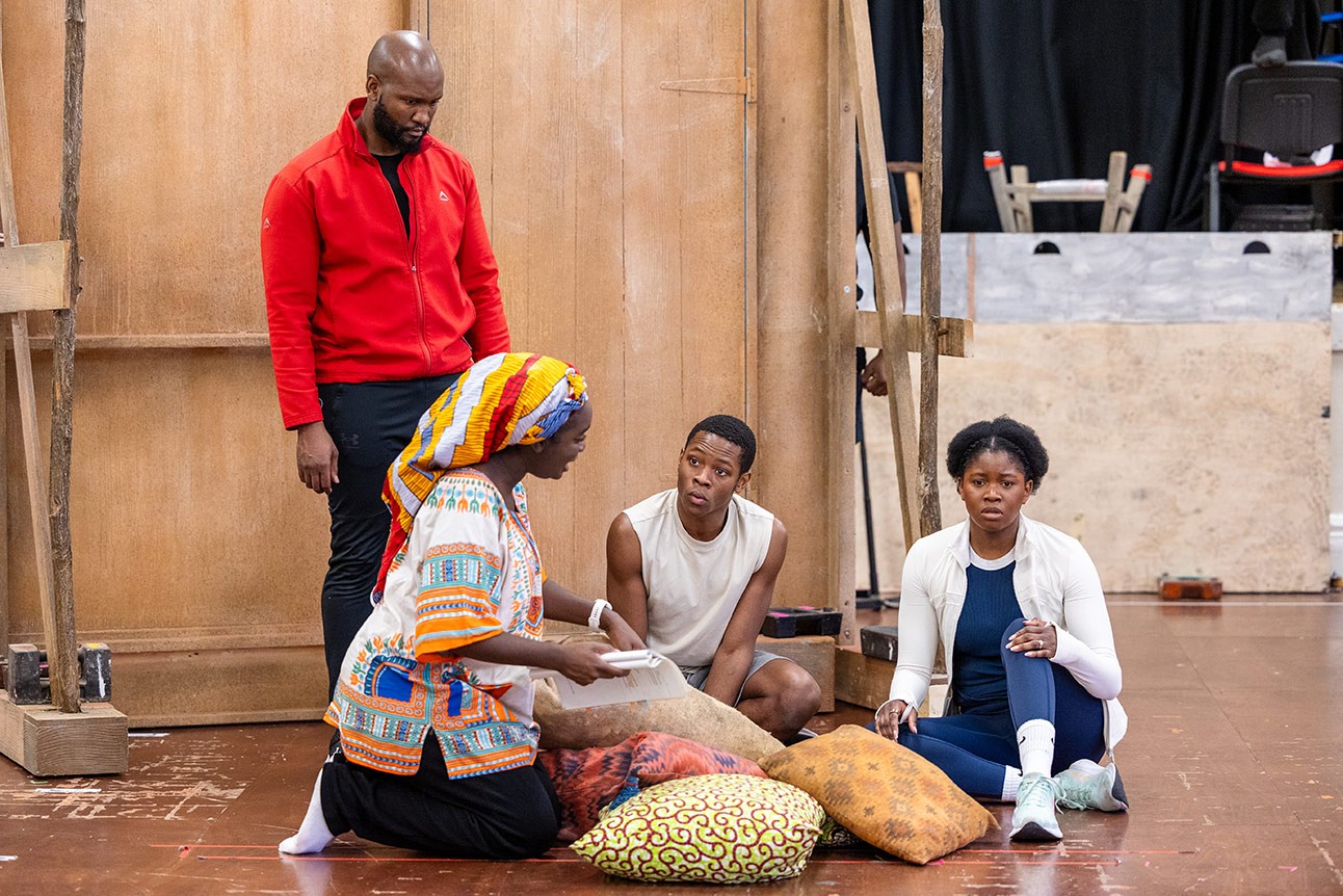 Four actors rehearse a scene from The Boy Who Harnessed the Wind, sitting and standing on a wooden stage.