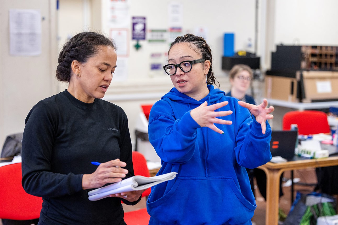 Two women in conversation during rehearsal, one taking notes, the other gesturing with hands, indoor setting.