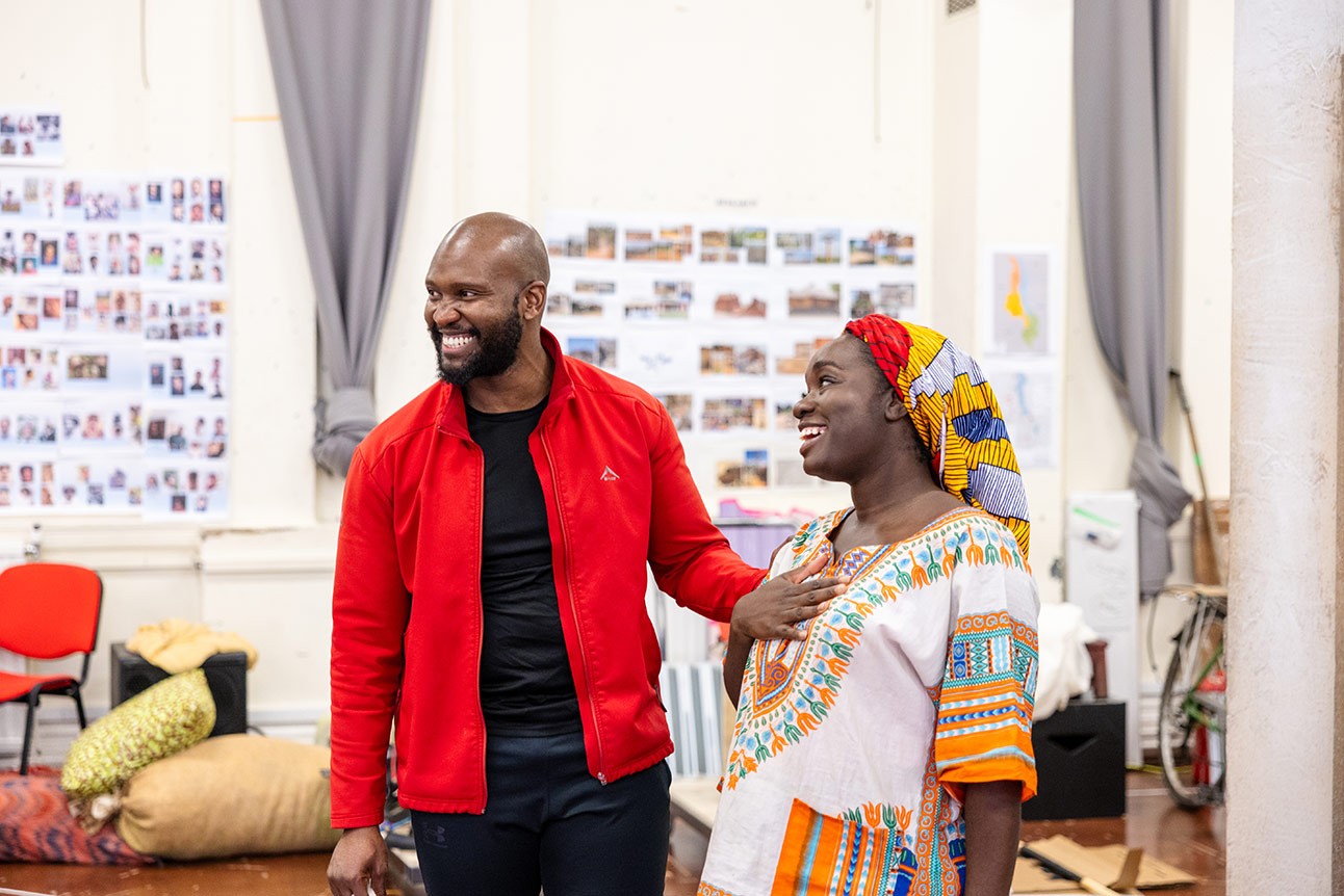 Two people, a man in a red jacket and a woman in colourful traditional clothing, smile in a rehearsal room.
