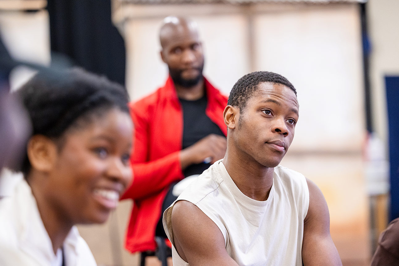Three actors rehearse, one in white sleeveless top foreground, another in red jacket blurred background.