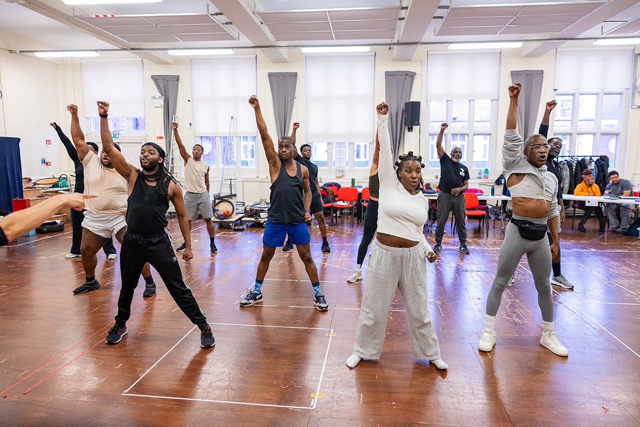 Actors rehearsing a dance routine in a spacious studio with wooden floors and large windows.