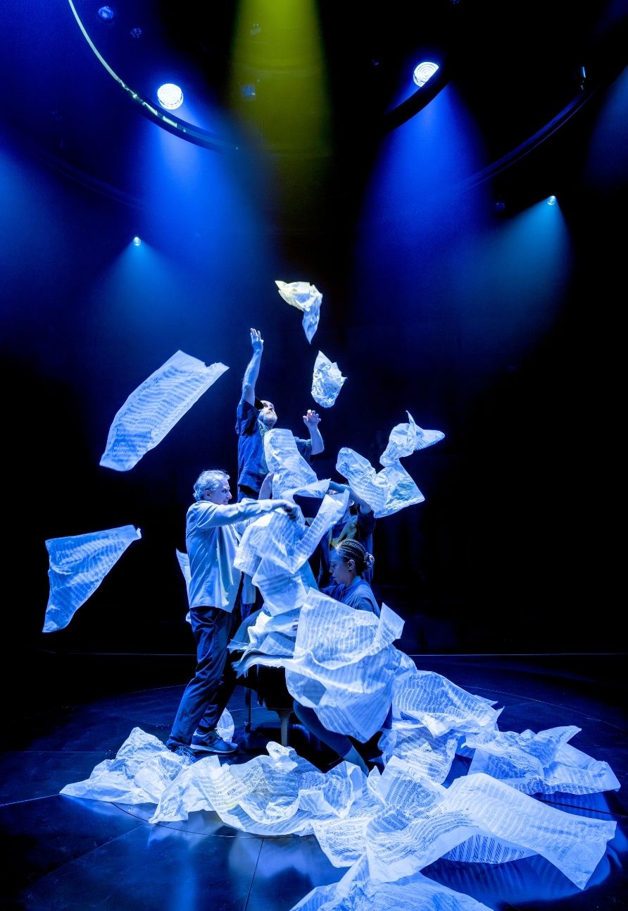 Performers under blue lighting throw white sheets of music paper into the air.