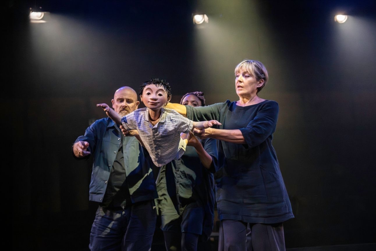 Performers on stage holding a puppet of a child, dramatic lighting, dark clothing, focused expressions, theatrical setting.