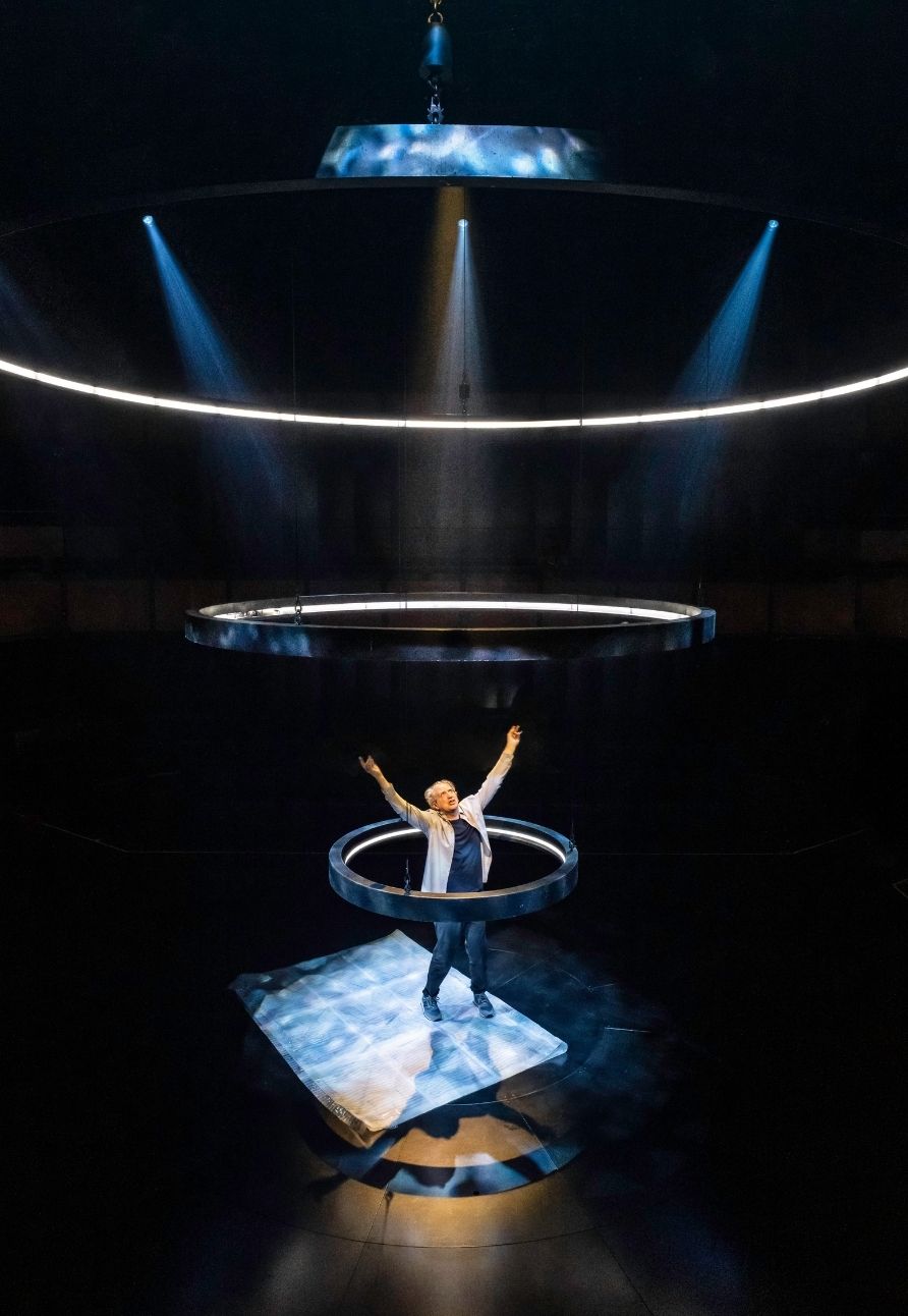 A performer stands with arms raised inside illuminated rings on a dark stage.