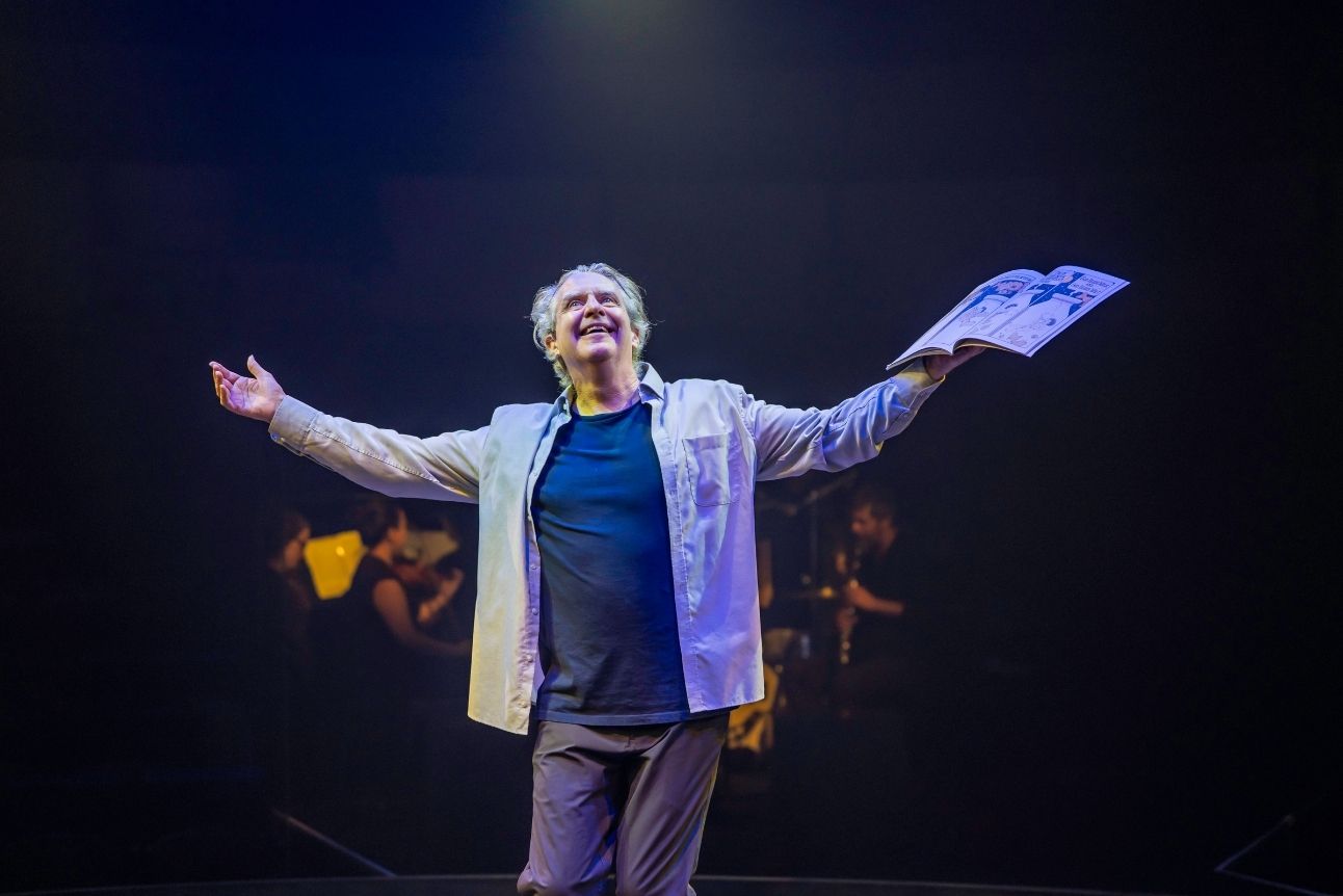 Phelim McDermott holds a children's story book, arms outstretched, on dimly lit stage.