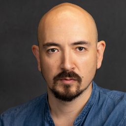 A man with a shaved head and goatee wearing a blue shirt against a dark background.