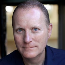 Dominic McChesney wearing a dark suit and blue tie, smiling against a plain grey background.