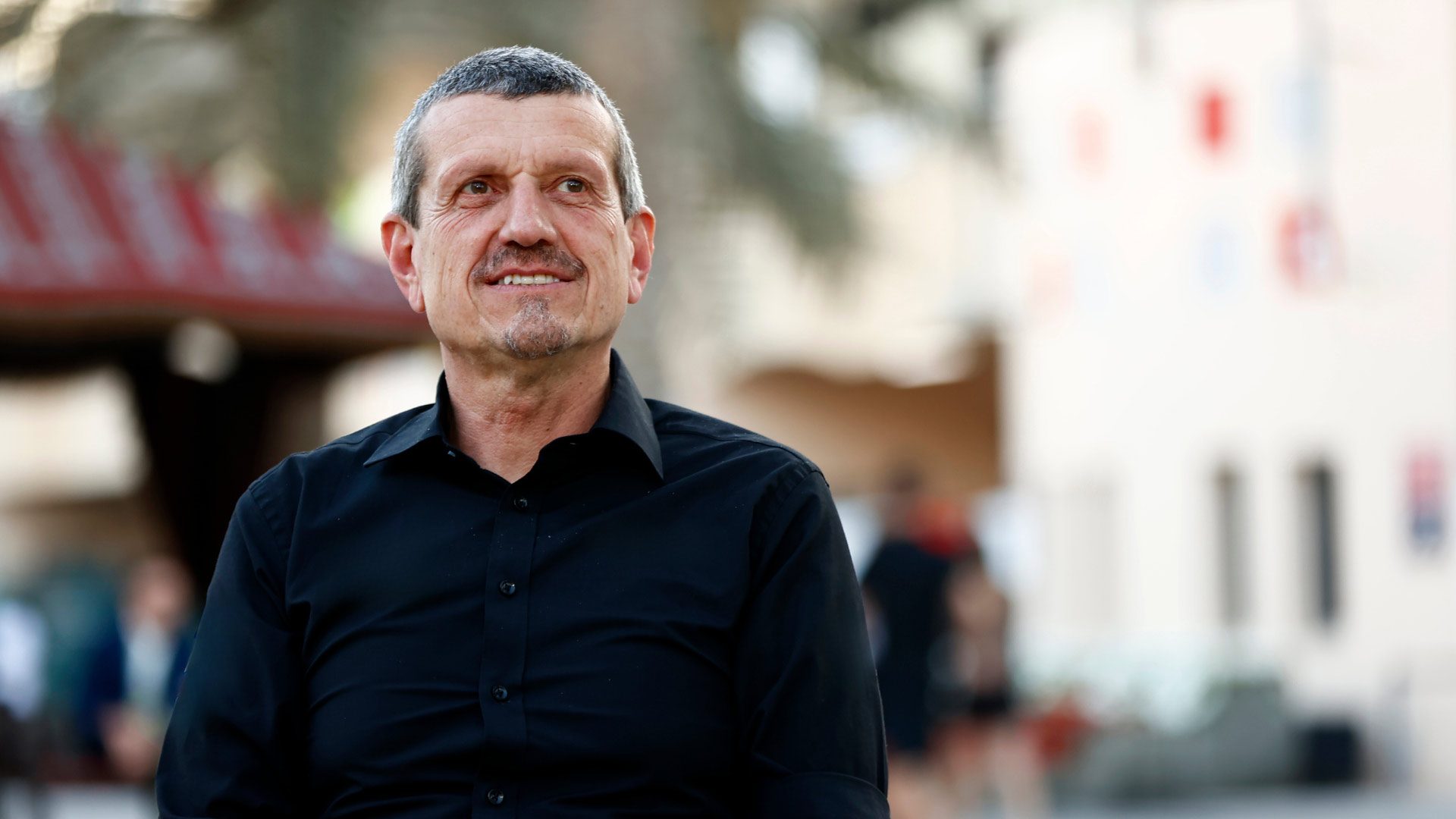 Guenther Steiner in a black shirt, looking slightly right, with a blurred outdoor background.