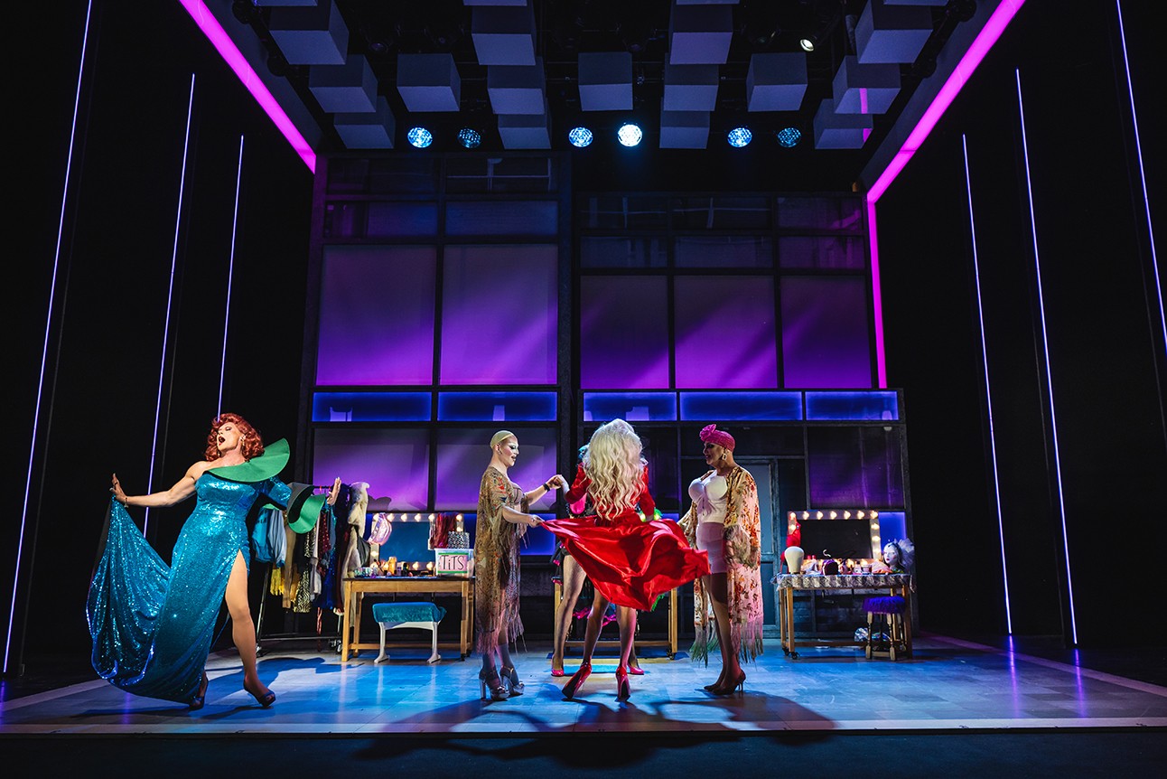 Three performers in colourful costumes pose on a stage with purple-lit backdrop and dressing tables with lights.