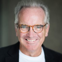 Jeremy Rose, smiling, wearing a dark blazer and light shirt, against a plain grey background.