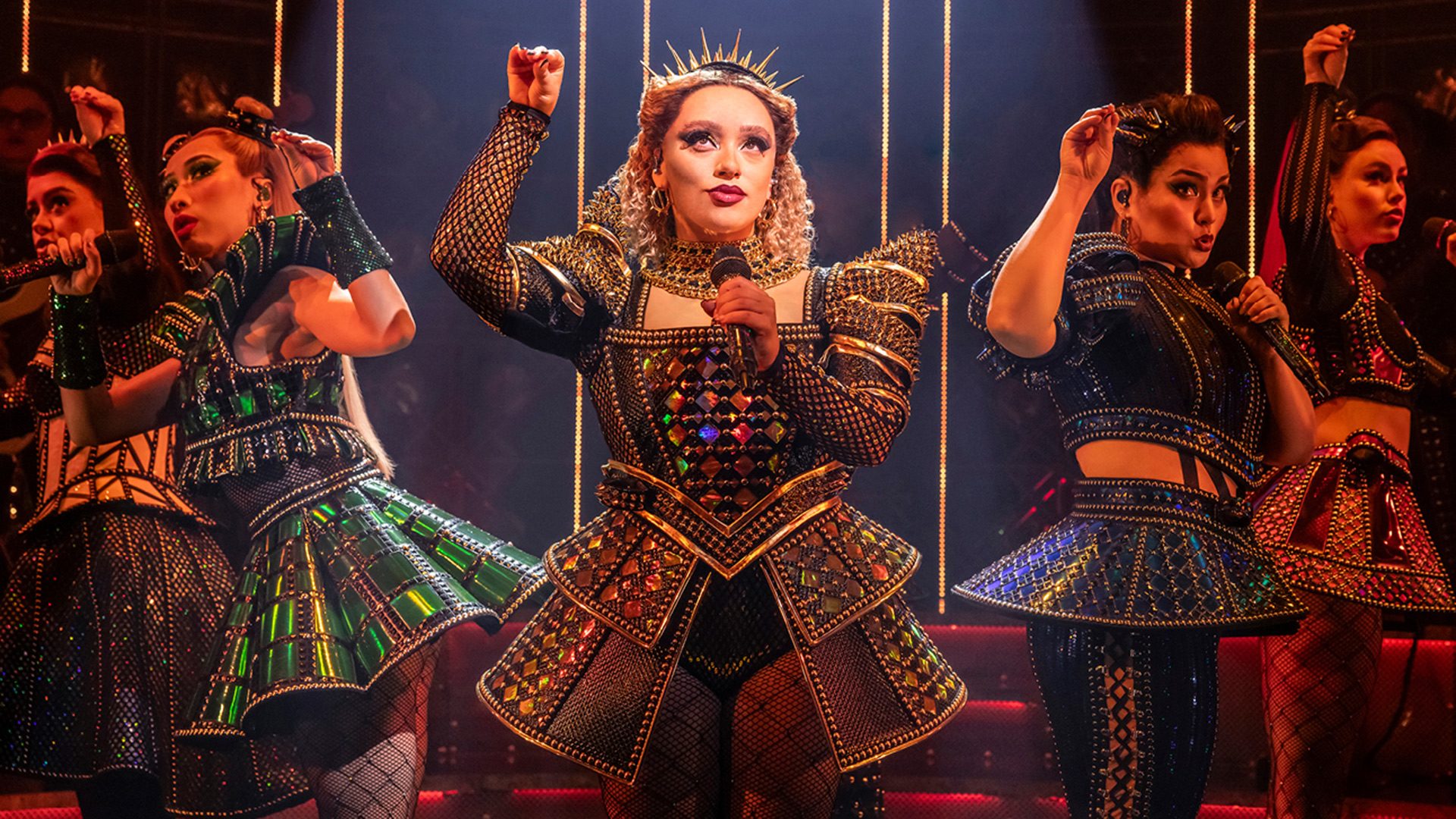 SIX Actress in ornate gold and black costume with crown, raising arms on stage with dark background.
