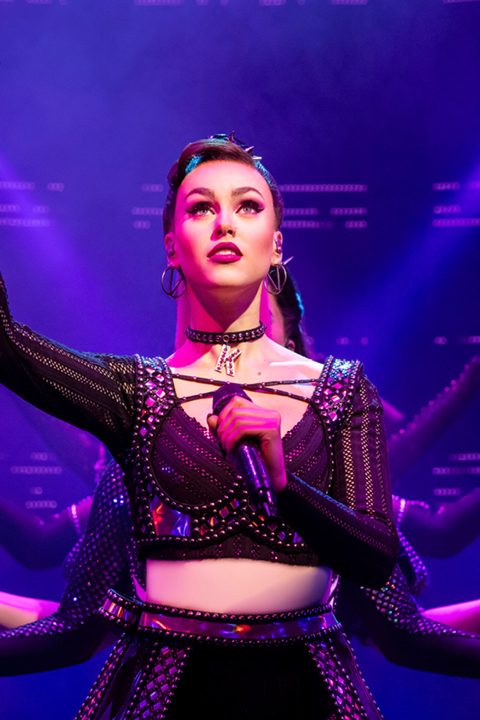 Female performer in black sequined outfit and choker, holding microphone under purple stage lighting.