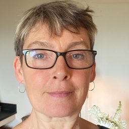 A woman with short grey hair and glasses, wearing hoop earrings, in a softly lit room.