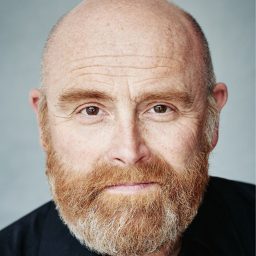 Bald man with a ginger beard, wearing a black shirt, against a grey background.